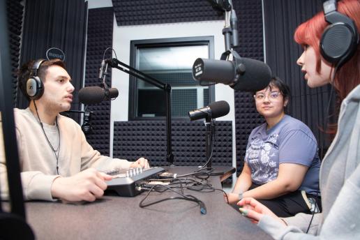 A man and 2 women collaborate in a radio studio, surrounded by microphones and sound equipment, engaged in conversation.